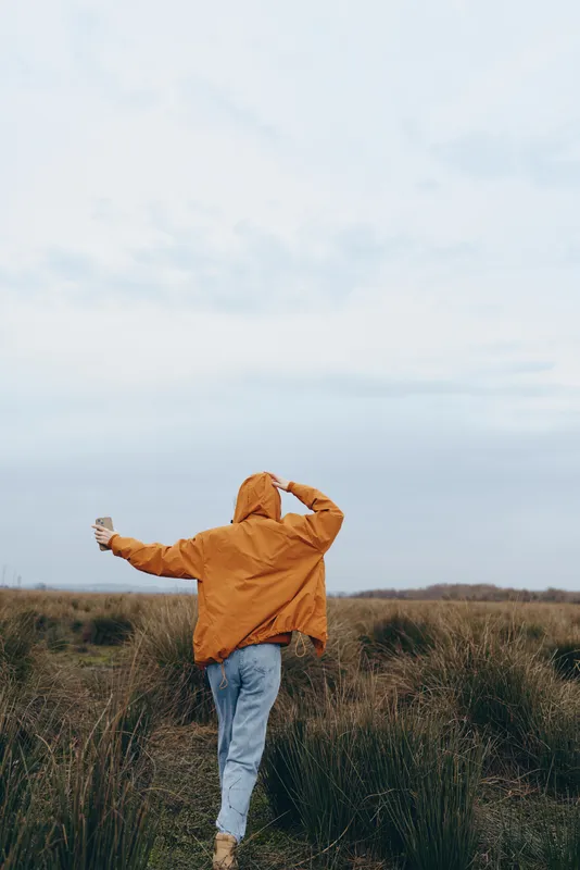 Woman lifestyle outdoors field running jacket jeans hood back view, arms open while moving in tall grass, orange coat on cloudy day, soft nature background, calm countryside