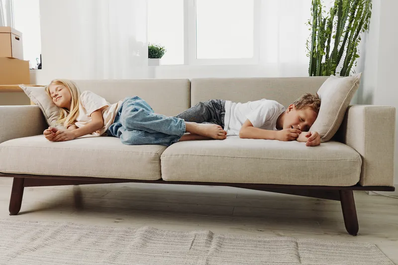 Two children lie relaxed on a beige sofa in a bright living room, enjoying a quiet moment. Casual home scene with natural light and cozy atmosphere.