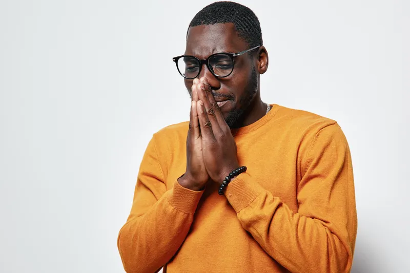 African American man with dark skin wearing glasses and orange sweater praying or meditating with closed eyes on white background. Thoughtful, spiritual, and peaceful moment.