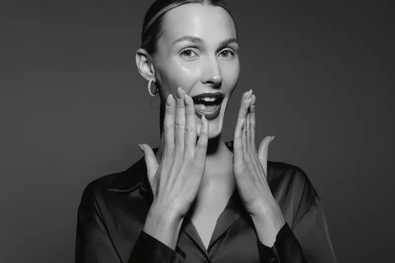 Excited young Caucasian woman with fair skin and slicked hair expresses surprise with open mouth and hands near face in black outfit. Concept of emotion and beauty.
