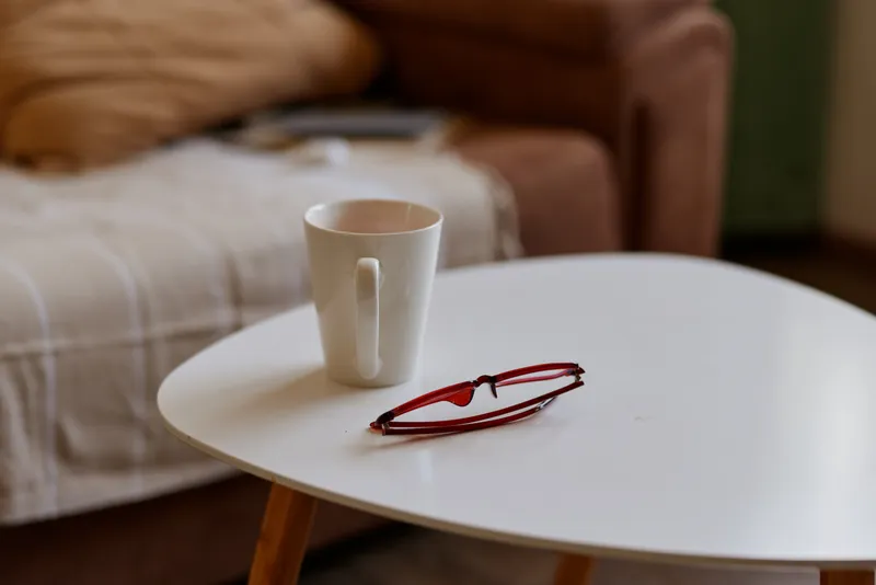 Cozy home lifestyle scene with a white table, mug and red glasses resting, soft sunlight and indoor calm, livingroom blurred backdrop, a simple morning moment