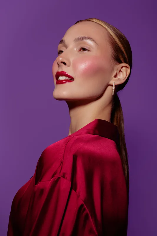 Portrait of confident woman with red lipstick and shiny skin wearing red satin blouse against purple background. Concept of beauty, fashion, elegance, and makeup in studio lighting.