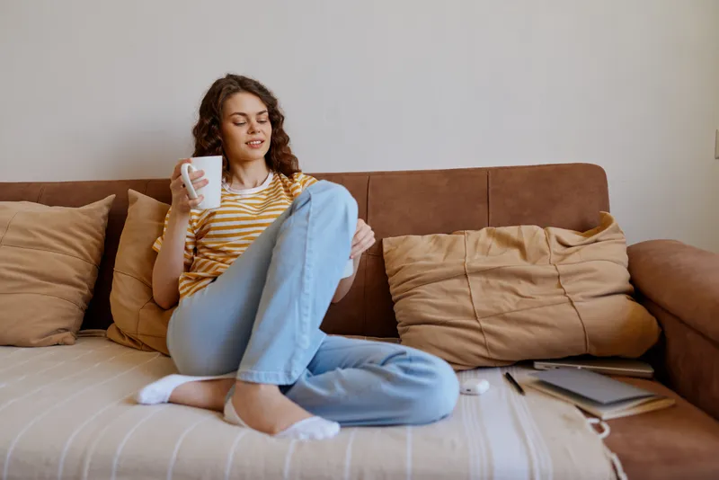 A relaxed woman sits on a couch at home, browsing on a smartphone, wearing a striped tee and jeans. Casual living room scene highlighting everyday comfort and digital connection.