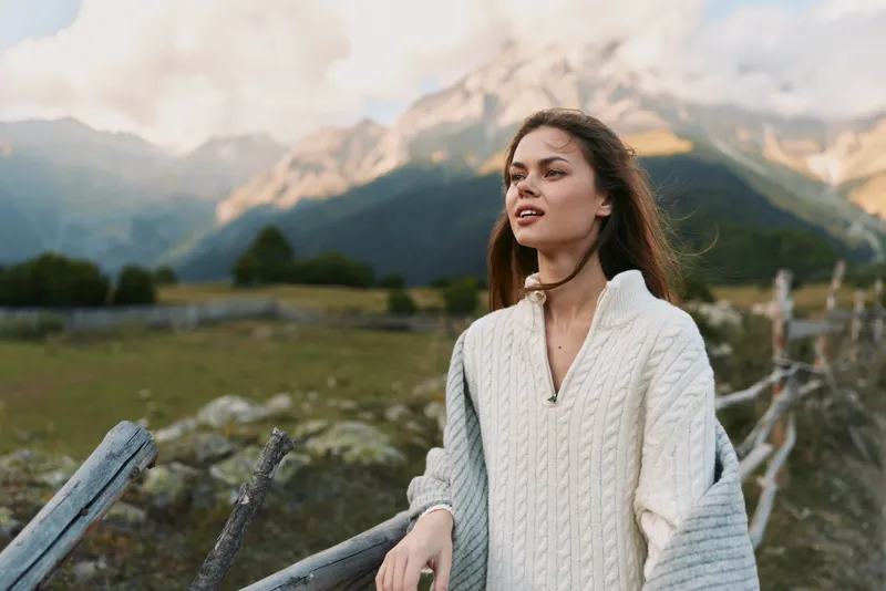 Woman in a chunky white knit sweater standing by a wooden fence in a mountainous landscape. Outdoor lifestyle shot conveys calm, freedom, and nature appreciation with soft natural light.