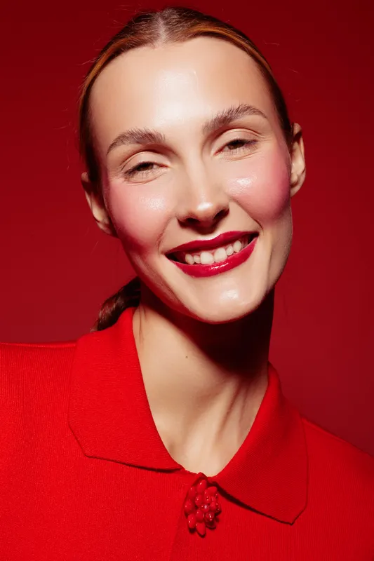 Smiling woman with red lipstick and red blouse poses against red background. Close-up portrait with natural skin texture and bright makeup. Concept of beauty and fashion.