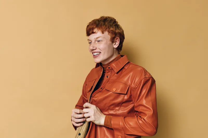 Young man with red hair wearing brown leather jacket smiles happily posing against beige background in studio. Casual style, fashion, emotion, portrait and lifestyle concept in warm tones.