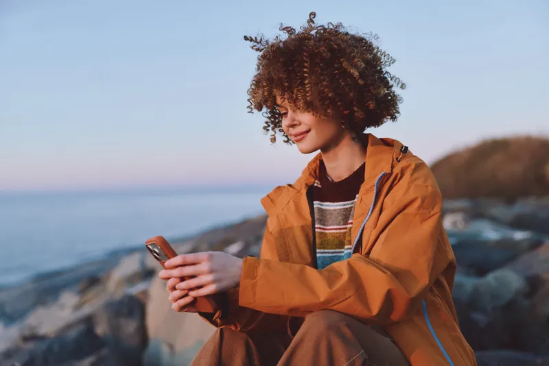 Woman lifestyle portrait outdoors by seaside, curly hair and orange jacket, smiling while using smartphone, ocean waves and rocky beach in soft focus, travel mood, morning light, fresh breeze