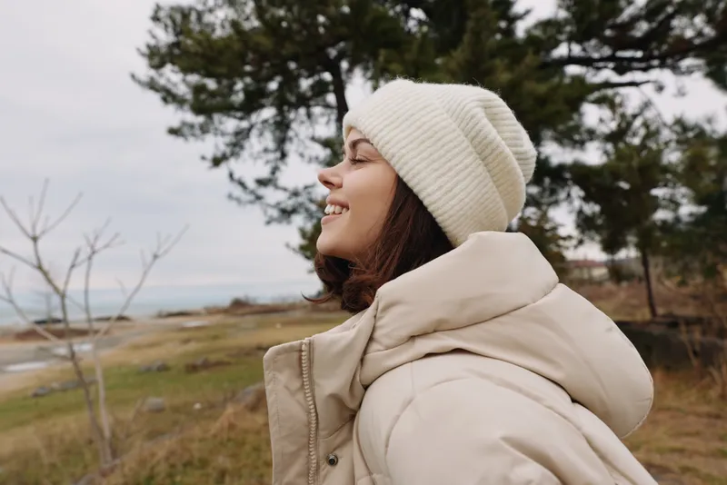 Woman enjoying outdoor winter scenery in a padded coat and cozy beanie, smiling as a coastal landscape with trees and open sky unfolds around her