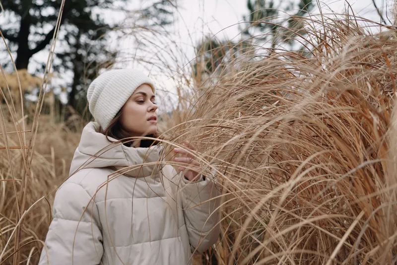 Winter, woman in a puffy coat and knit hat stands amid tall dried grasses in a quiet field, capturing serene outdoor moments, soft textures, and calm natural scenery.