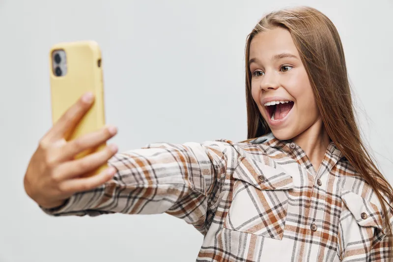 Girl with long hair wearing checked shirt taking selfie with smartphone smiling and looking excited on neutral background in studio lighting.