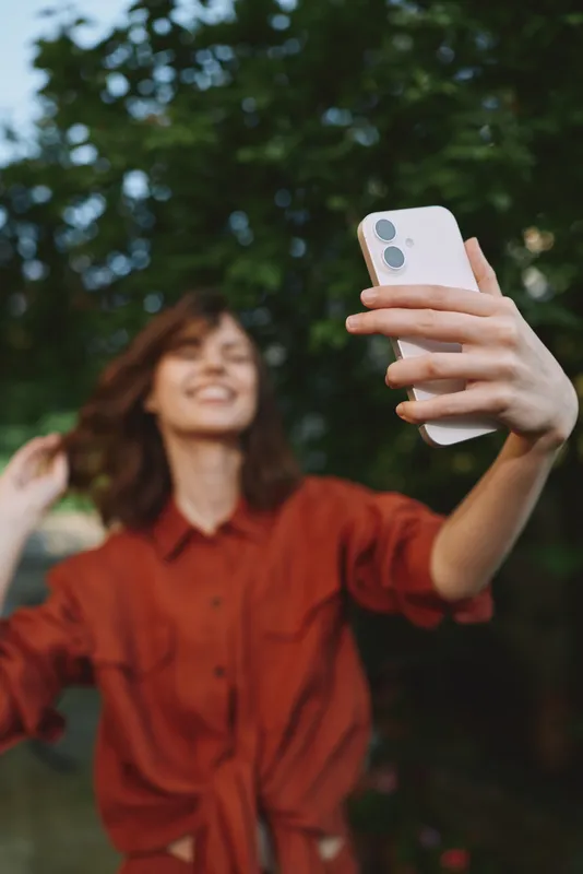 Smiling woman in a red shirt taking a selfie outdoors, using a smartphone. Candid lifestyle shot capturing joy, connection, and casual fashion in a green park setting