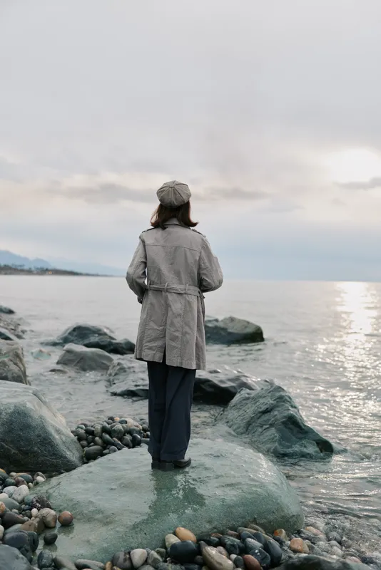 Woman in grey coat and hat stands on rocky shore, facing calm sea and cloudy sky. Concept of solitude, contemplation, and peaceful nature moment outdoors by water.
