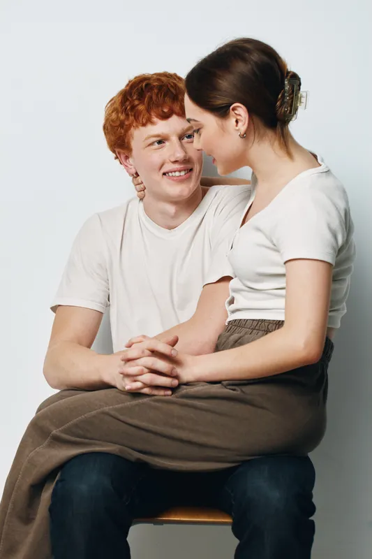 Young couple sitting close together holding hands looking at each other, smiling and happy with casual clothes on white background.