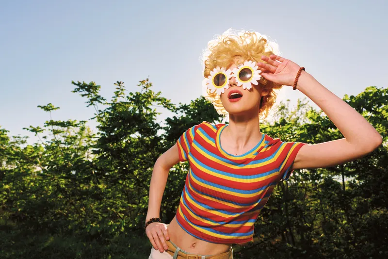 A playful woman outdoors wears a striped top and flower sunglasses, posing with whimsy. Bright retro film color enhances the vibrant, carefree mood and fun style for fashion lifestyle shots