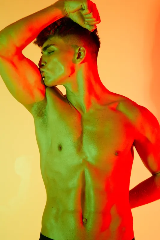 Young muscular man posing shirtless and flexing bicep with colorful red and green light effect on skin against soft yellow background while showing strength and fitness.