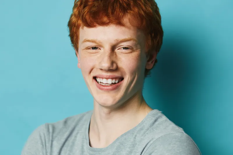 Young man with red hair and freckles smiling and looking at camera against blue background. Closeup portrait of happy male with casual shirt and friendly expression.