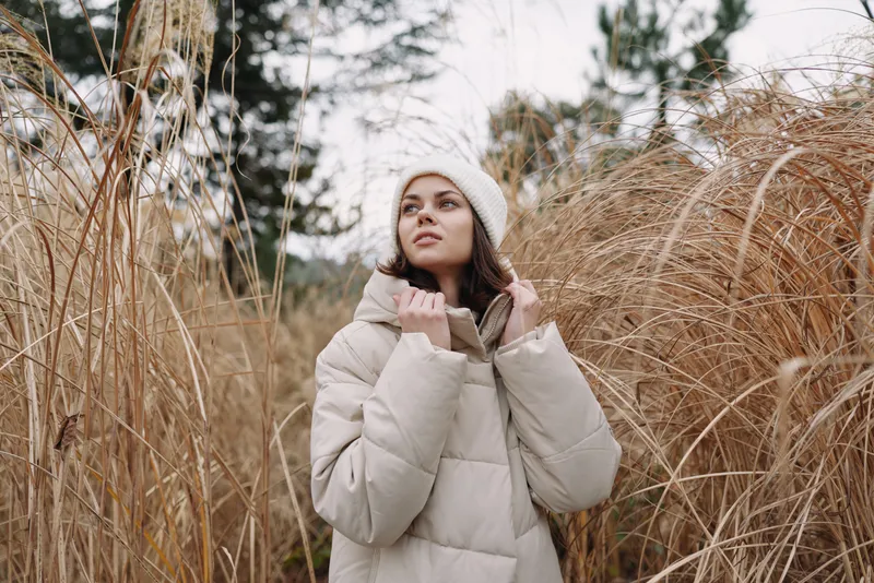 Winter coat, woman and outdoor portrait among tall grass in a pale field, she gazes upward with a soft expression, bundled in a cozy beige jacket and beanie.