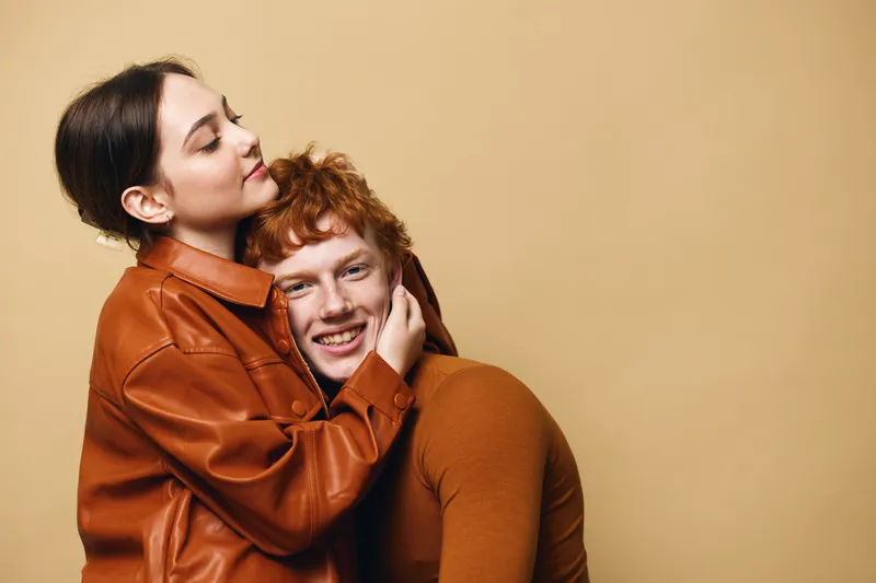 Couple hugging with love and affection, young woman embracing smiling man in casual brown clothing with warm beige background, relationship and happiness concept.