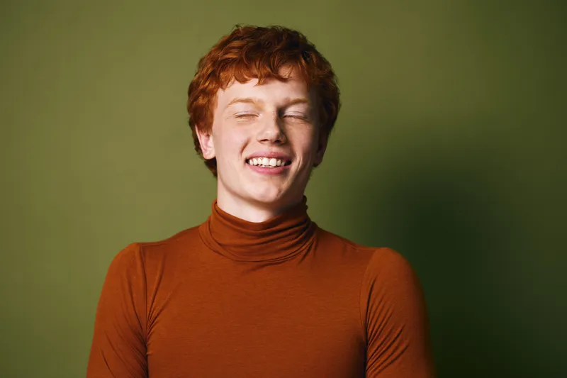 Young redhead woman with freckles smiling with closed eyes wearing brown turtleneck sweater on green background. Portrait of cheerful female person with short hair and natural look.