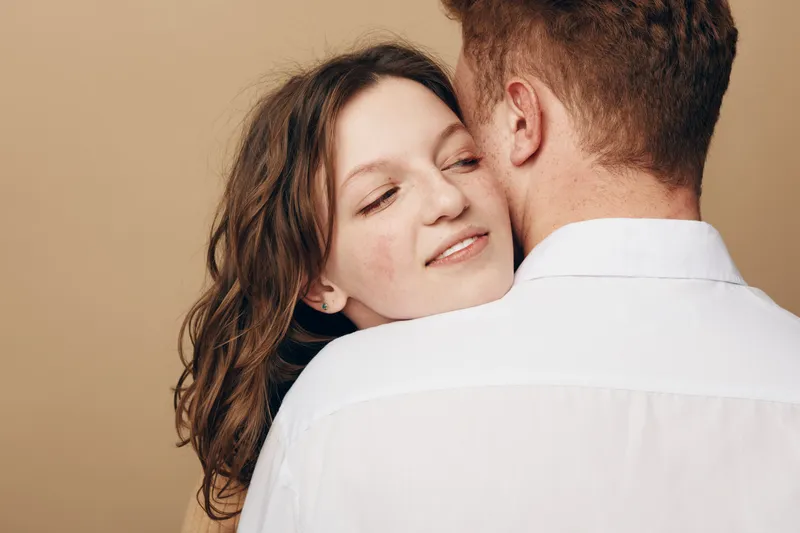 Young couple embraces tenderly, woman resting her head on man’s shoulder with closed eyes. Intimate moment of affection and love captured in soft natural light on neutral background.