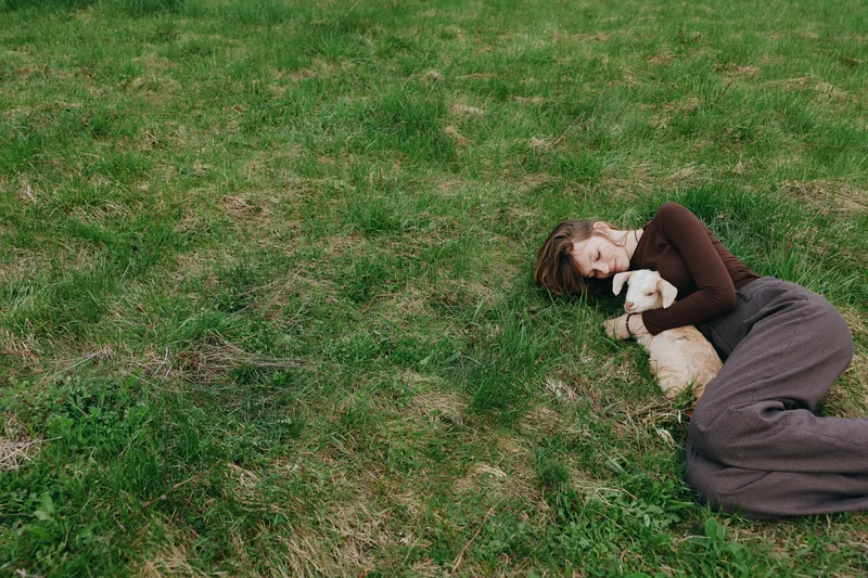 A woman lies on the grass embracing a goat kid in a peaceful outdoor setting. Warm daylight, relaxed mood, rustic atmosphere, gentle companionship, nature connection, intimate moment in natural light.