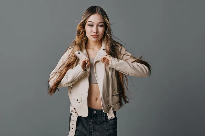 Young woman with long hair wearing beige jacket and dark jeans posing on neutral background with confident expression and natural makeup in studio lighting.