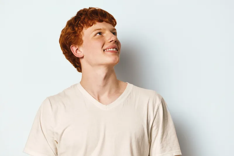Young man with red hair wearing plain light shirt looking up with smile on light background. Portrait of male model with fair skin and curly hair in casual clothing indoors.