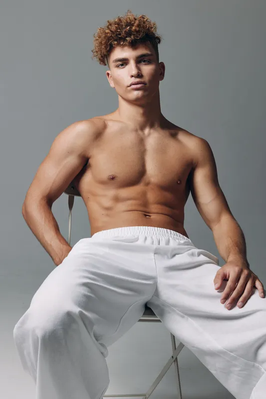 Young man with curly hair sitting on chair shirtless wearing white pants against gray background. Muscular body, confident pose and casual style in studio portrait photography.