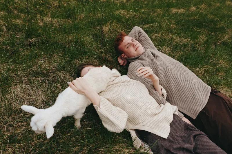 Two people lie on the grass with a white goat kid beside them, sharing a calm outdoor moment. Cozy sweaters, warm light, and natural surroundings convey lifestyle, companionship, wellness, and simple