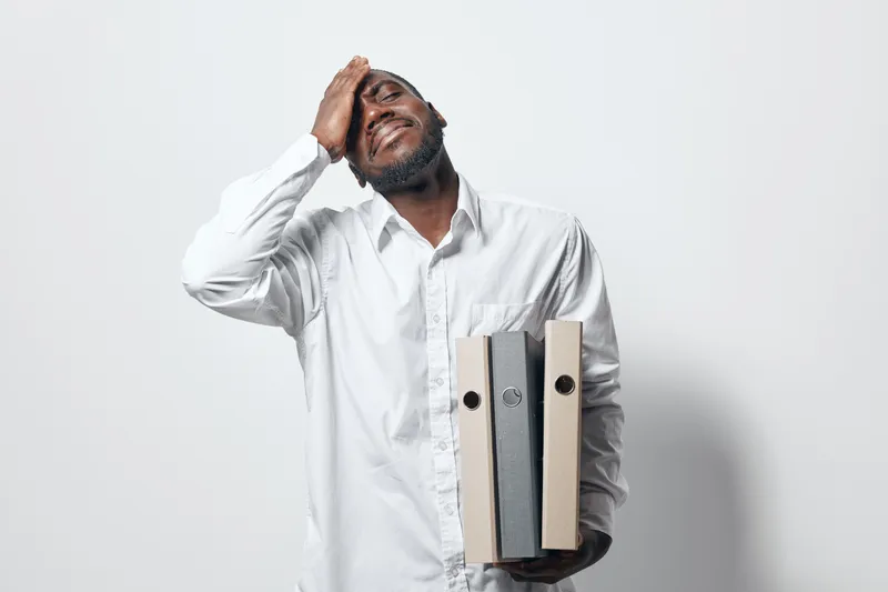 African American man in white shirt holding folders and touching head with tired expression on white background for stress and work pressure concept.