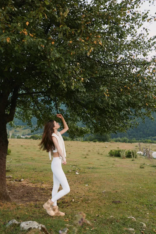 Cheerful woman outdoors, standing in a sunlit field under a leafy tree, reaching up with relaxed pose, white outfit, natural colors, carefree mood