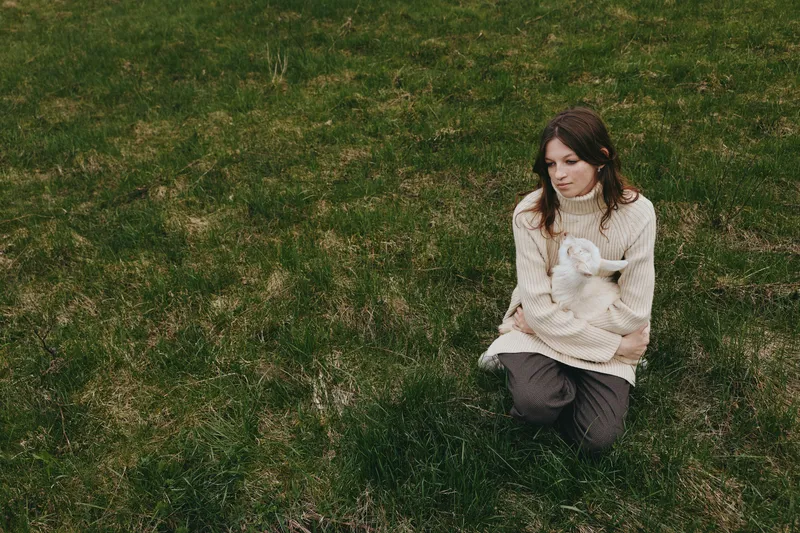 Woman kneels on grass, gently cradling a goat kid in her arms. Natural light highlights a peaceful outdoor moment, emphasizing connection, care, and a simple lifestyle with animals.