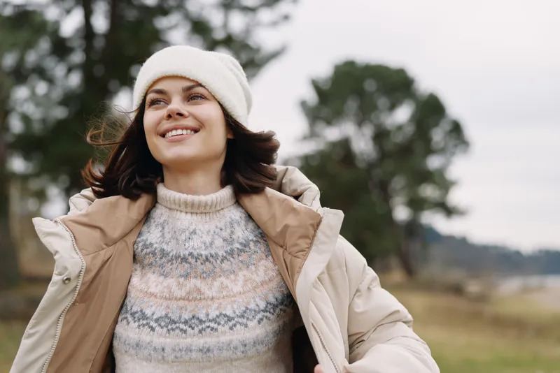 Woman outdoors wearing a cozy coat and knitted sweater, smiling and relaxed as wind moves her hair, capturing casual winter fashion and a carefree lifestyle in a breezy landscape.