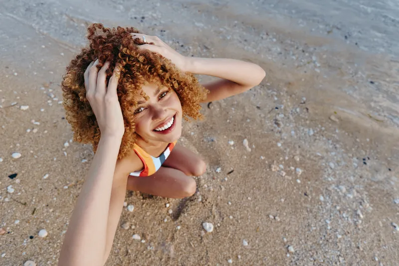Smiling woman with curly hair lies on beach sand, hands behind head, sunlit outdoor portrait, relaxation and summer vacation mood, carefree joy, concept of leisure, wellbeing and mental health