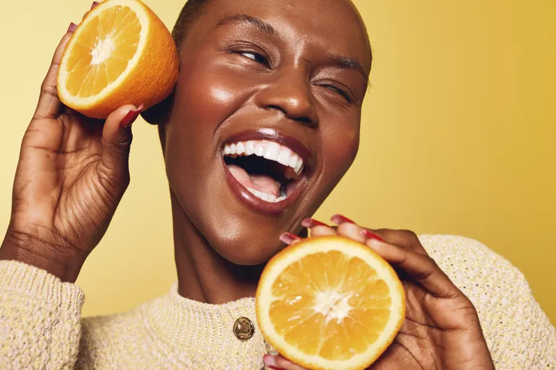 Happy African woman with dark skin holds fresh orange halves near face smiling with joy. Bright yellow background and cozy beige sweater create warm and cheerful mood. Concept of health and vitality.