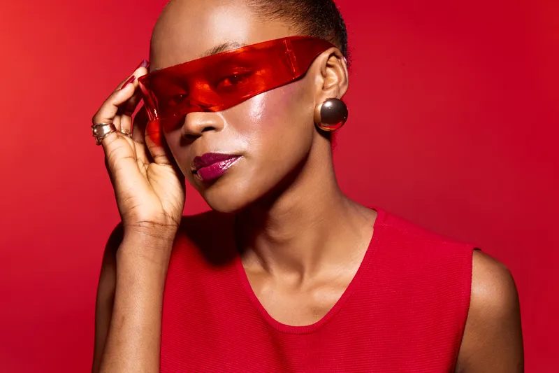 Stylish dark skinned African American woman wearing red futuristic sunglasses and red sleeveless top poses confidently on red background. Concept of fashion, beauty, and modern style.