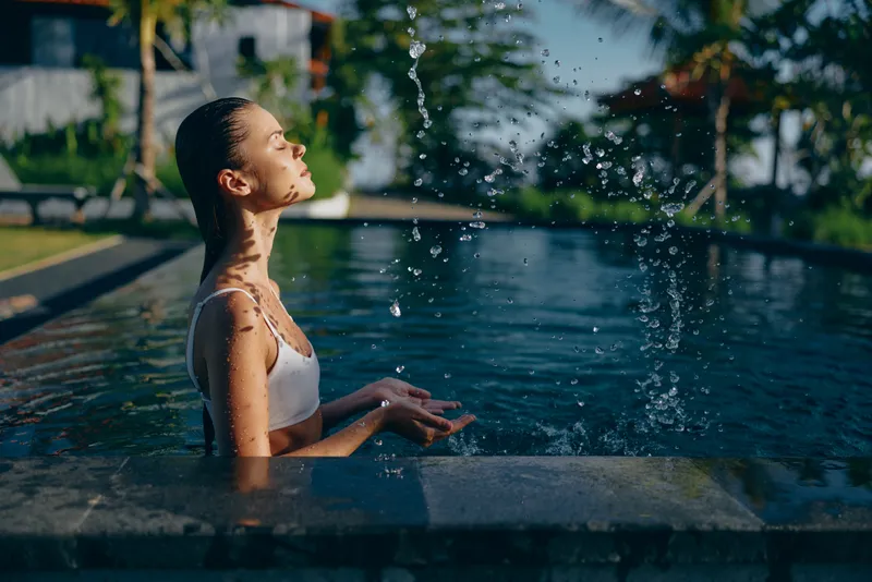 Young woman with long wet hair relaxes in an outdoor pool while water splashes around her under natural sunlight, enjoying a peaceful moment of mindfulness and wellness.