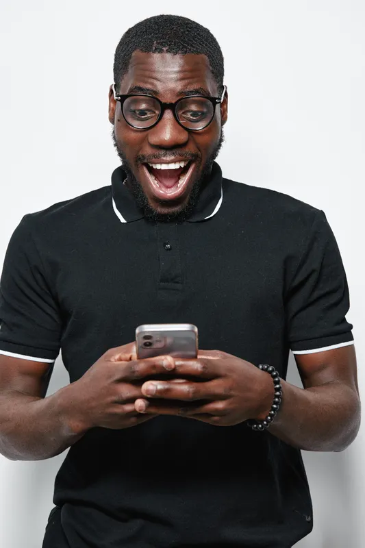Black man with dark skin and glasses wearing black polo shirt smiling excitedly while looking at smartphone in hands on white background.