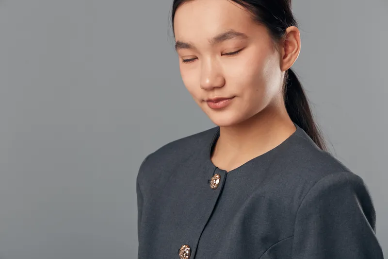 Young woman with closed eyes and calm expression wearing dark blazer, standing in front of gray background, natural light highlighting face and professional attire with neat hairstyle.