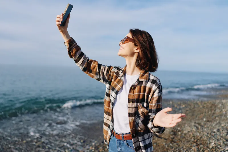 Woman on a rocky beach taking a selfie with phone, wearing plaid shirt and sunglasses, open arms under a sunny sky, enjoying a coastal day and carefree travel mood
