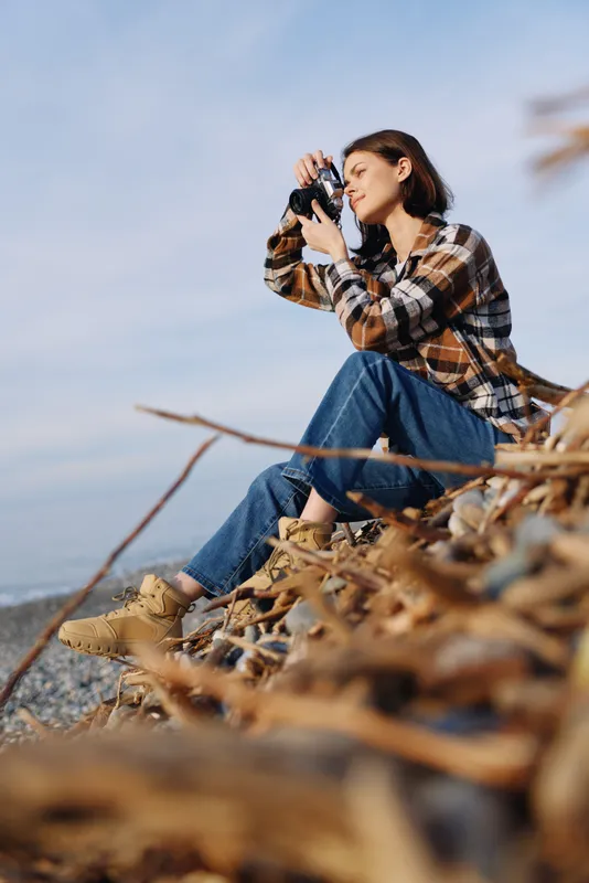 Female photographer sits on a rocky shore with a camera to her eye, wearing a plaid shirt and jeans, focused on capturing coastal scenery as dry grasses frame the foreground.