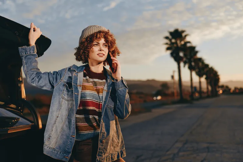 woman, lifestyle, street, car, denim, jacket, outdoor, sunset, road, curly hair, striped sweater, leaning near car door, one hand raised, relaxed smile, urban evening travel portrait