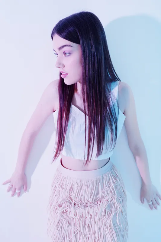 Young woman with long dark hair wearing a white top and fringed skirt stands against a light background. Concept of beauty, fashion, and modern style with soft lighting.