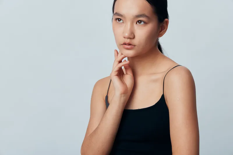Young woman with natural skin and black tank top posing thoughtful on plain light background. Female model looking sideways with calm expression and minimal makeup in studio.