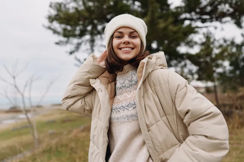 Woman posing outdoors in a warm beige winter coat, smiling confidently in a chilly countryside scene with a knit sweater and hat, suggesting casual fashion, fresh air, and outdoor lifestyle.