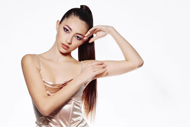 Portrait of a young woman with sleek ponytail and creative vibrant makeup posing with graceful arms against white background. Concept for fashion beauty or artistic style.