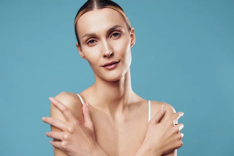 A young woman with blonde hair stands against a blue backdrop, touching her shoulders in a calm studio portrait, conveying self care and quiet confidence.
