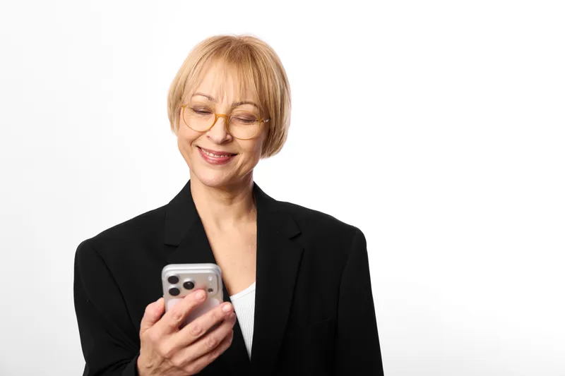 Smiling elderly woman wearing glasses holding smartphone, business portrait in studio, white background, communication and technology for everyday life, calm mood
