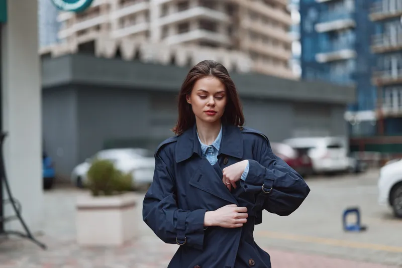 A young woman in a navy trench coat adjusts her sleeve on a bustling city street, exuding confident urban style, a focused gaze, and a self-assured mood.