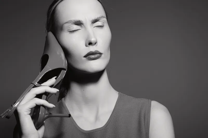 Caucasian woman with light skin holds a high heel shoe against her face with closed eyes, expressing discomfort or pain in black and white photo.